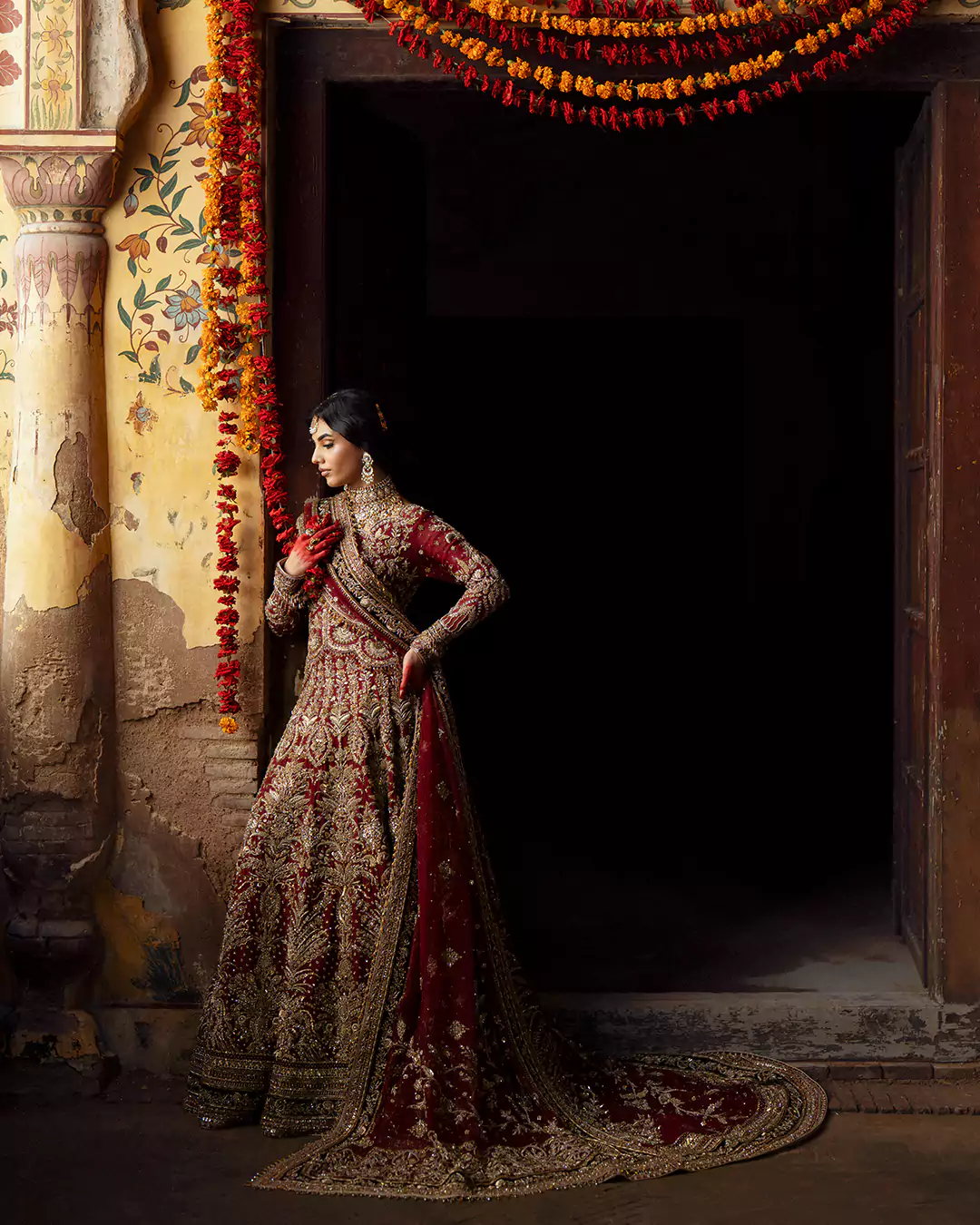 Model wearing a deep red heavily embellished bridal lehenga with a trailing veil alongside marigold garlands.