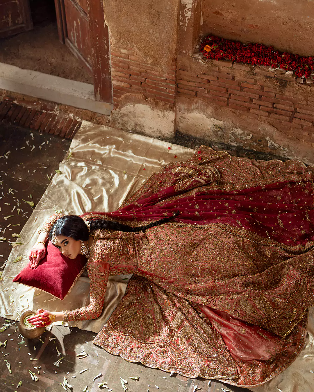High-angle shot of a model lying on satin fabric wearing a gold and red heavily embroidered bridal attire.