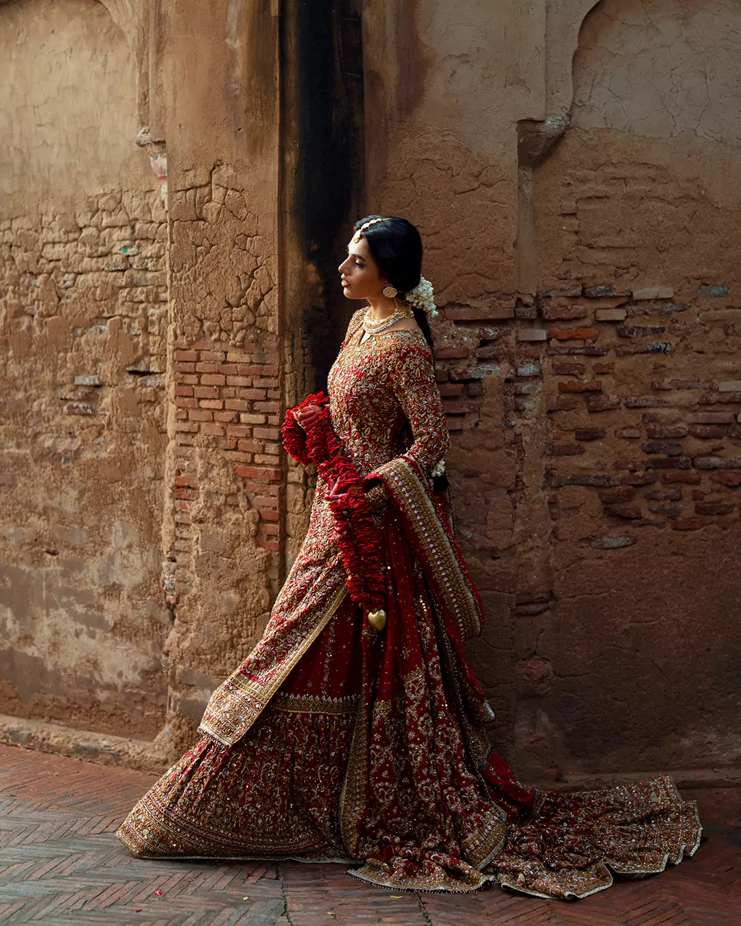 Full profile view of a model in a deep red zardozi-embroidered bridal ensemble holding a red rose garland.