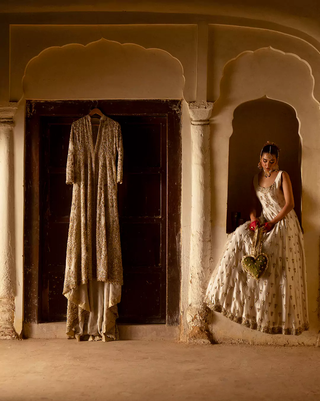 Model seated in a niche wearing a sleeveless ivory lehenga with an embroidered jacket hanging on a wooden door.