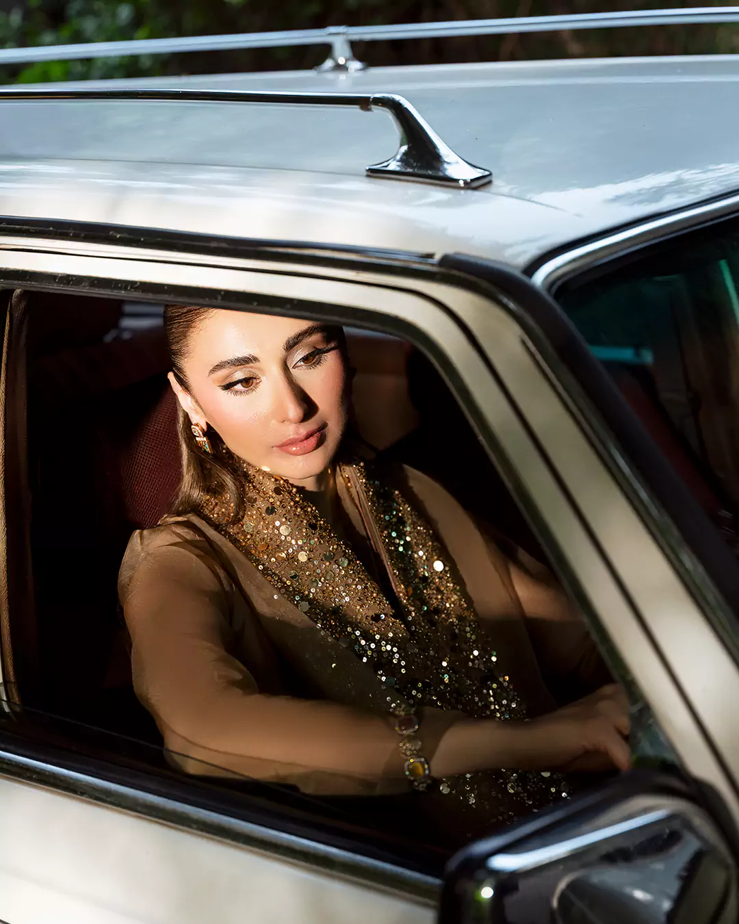Model wearing a brown outfit while seated in the driver's seat of a vintage car for the AKKS by Attya Luxury Pret campaign.