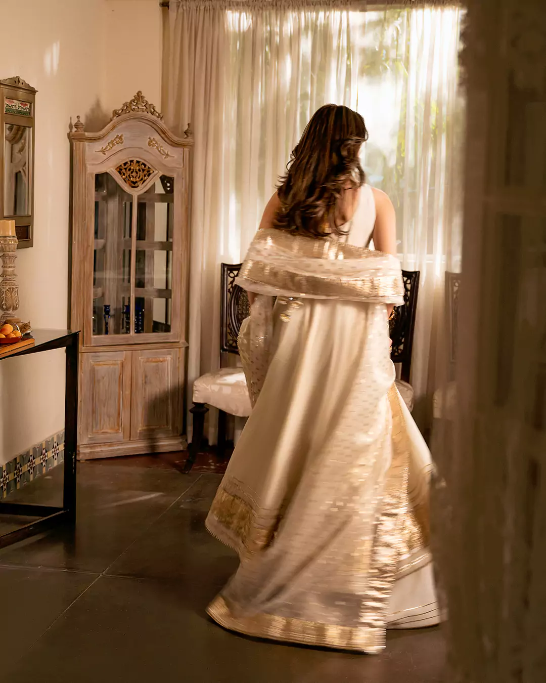 Rear-view fashion photograph of a model showcasing an off-white outfit in a classic drawing room for the AKKS by Attya campaign.