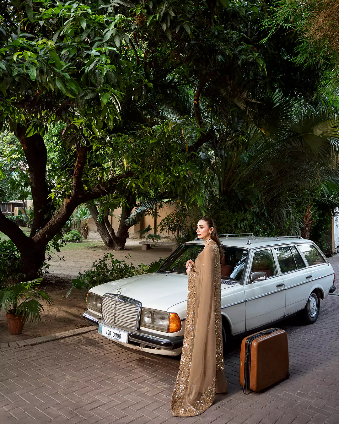 Model wearing a brown outfit and holding a brown suitcase beside a vintage Mercedes-Benz for the AKKS by Attya campaign.