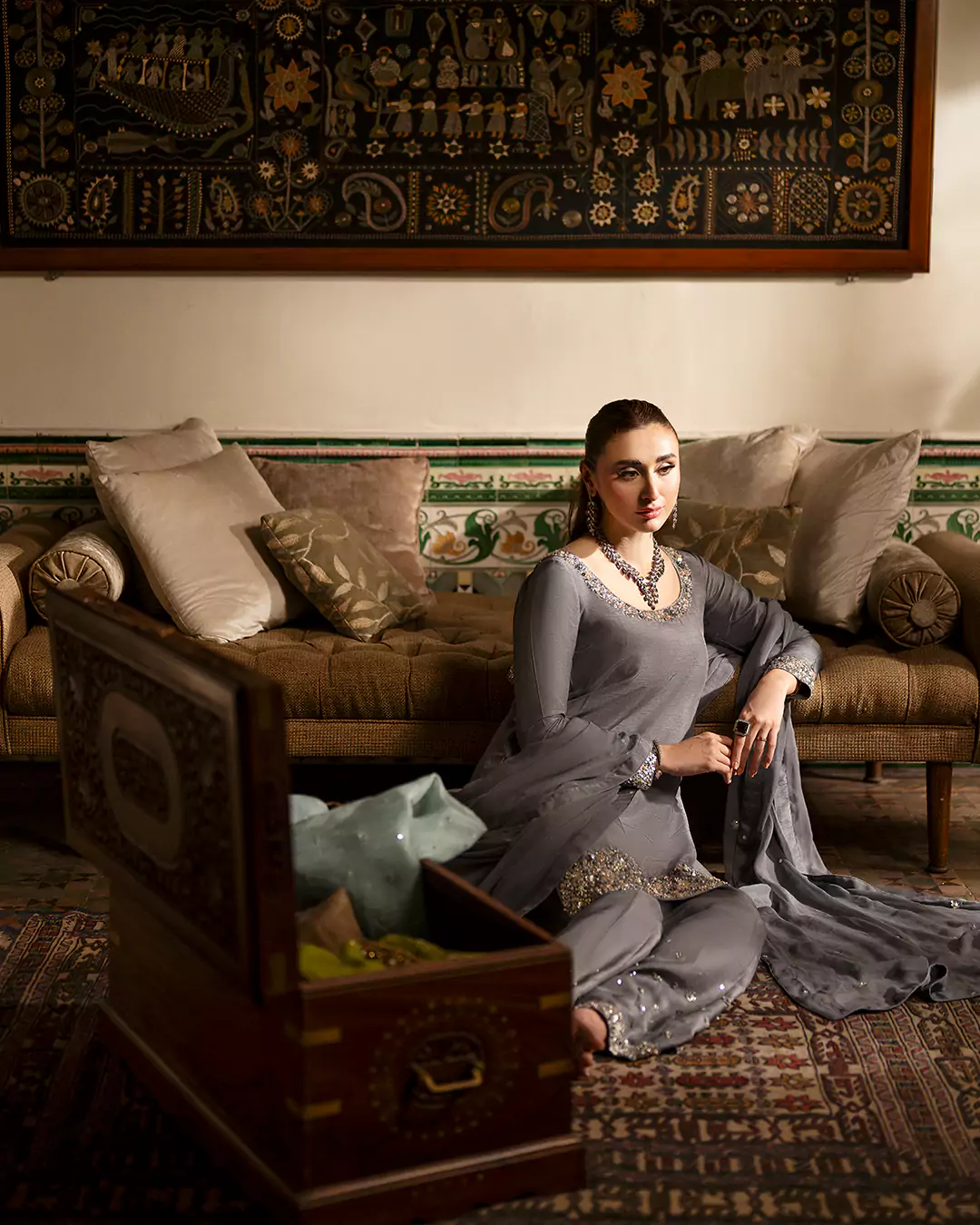 Model wearing a metallic-grey outfit, seated on the floor beside a sofa with one hand resting on it and a brown wooden chest in front.